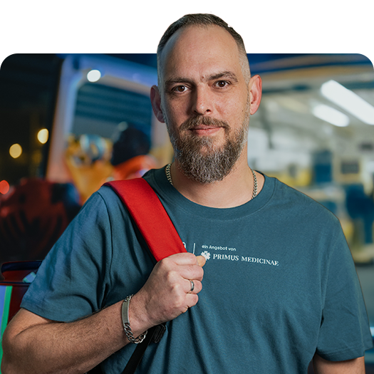 Patrick Schmude vor Rettungswagen mit Primus Medicinae-Shirt und Notfallrucksack über einer Schulter