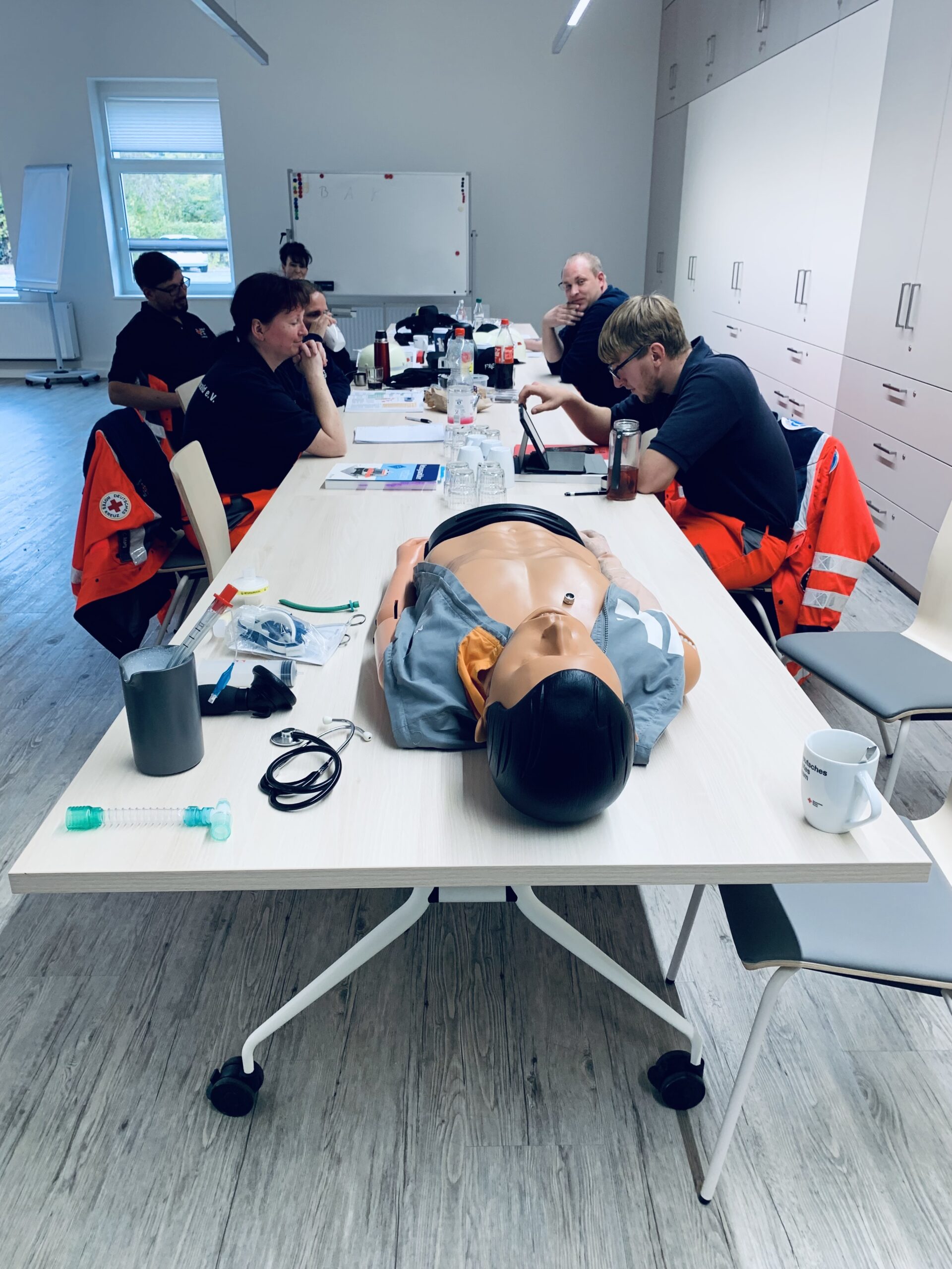 Mehrere Personen mit Rettungssanitäterjacken sitzen an einem großen Tisch in einem Übungsraum. Auf dem Tisch befindet sich Schreibzeug, ein Dummy sowie Flaschen und Gläser.