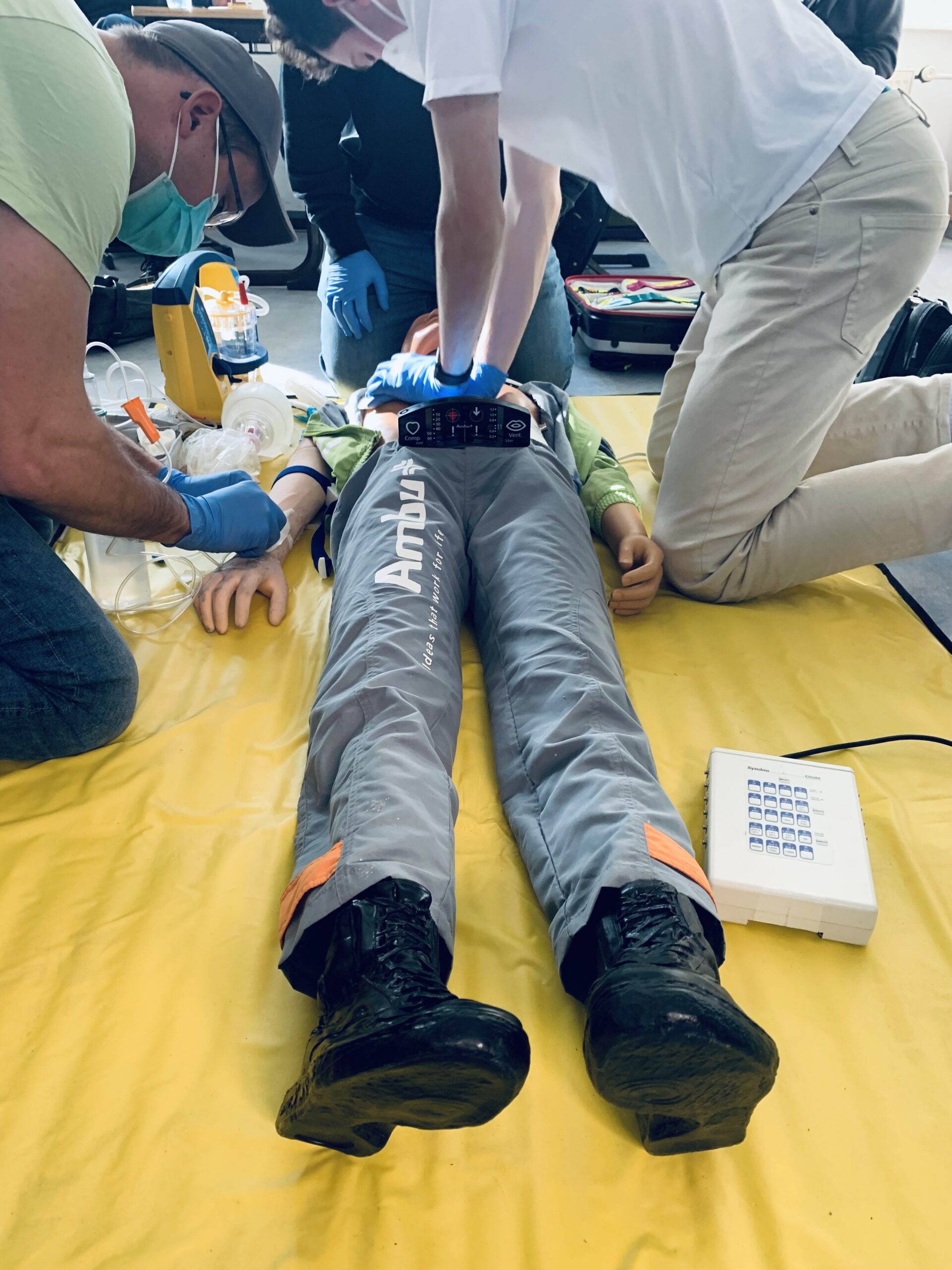 ein Dummy-Patient liegt auf dem Boden, eine Person übt Herzlungenmassage, eine andere legt einen Zugang. Beide tragen Mundschutz. Im Hintergrund kniet eine weitere Person und es liegt Notfallausrüstung herum.
