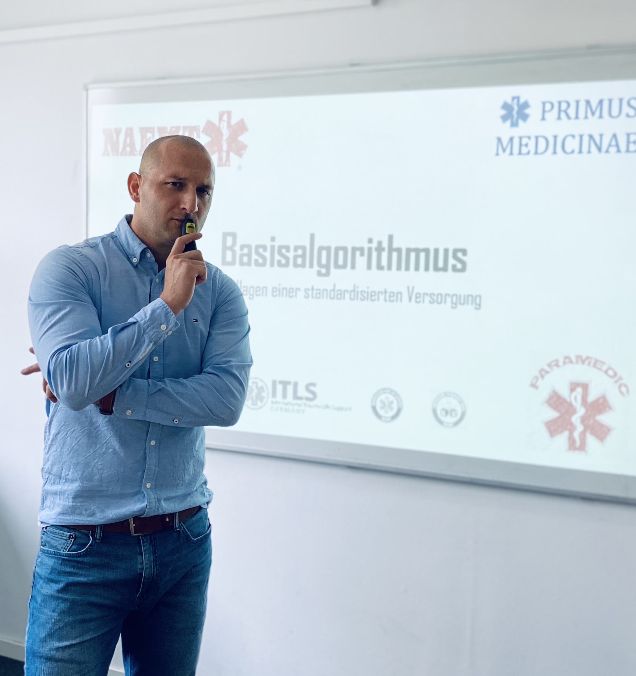 Boris Schweizer in Hemd und Jeans schaut den Betrachter kritisch an. Im Hintergrund ist eine Leinwand, auf der man Inhalte des Rettungssanitäterkurses sieht.