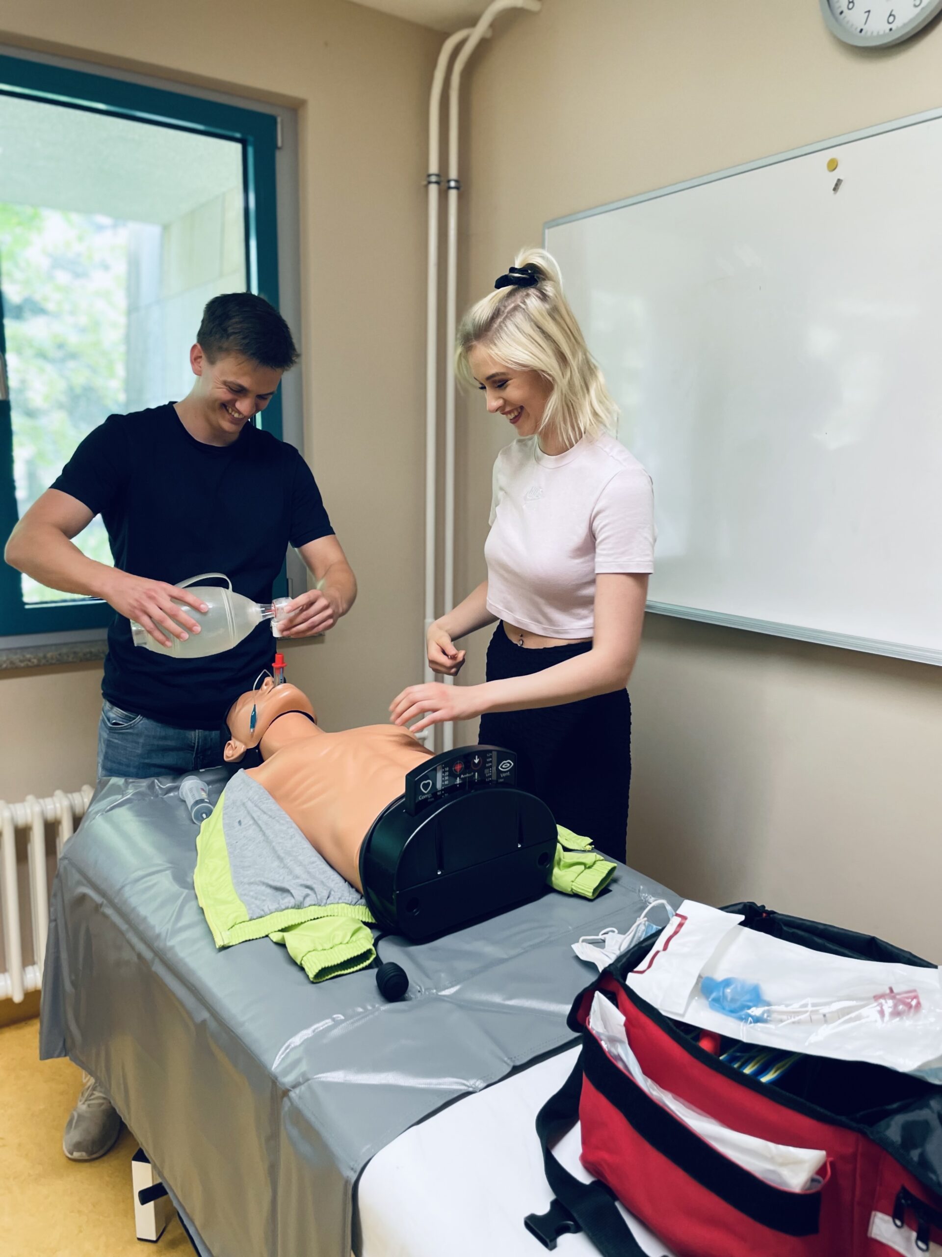 Zwei lächelnde Auszubildende (ein junger Mann, eine junge Frau) üben den Einsatz des Beatmungsbeutels an einem Dummy, der auf einer Liege liegt.