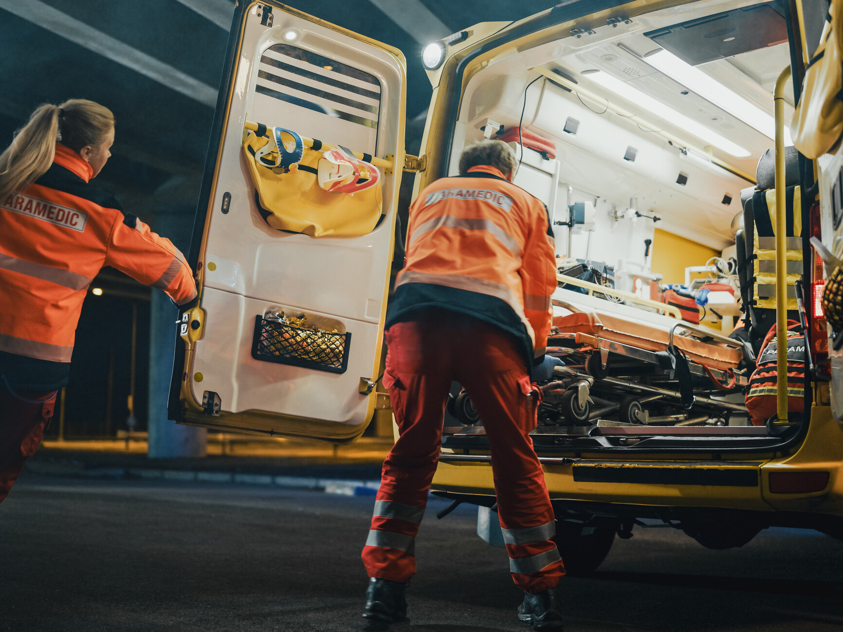 Ein Rettungssanitäter schiebt eine Person auf der Trage in den offenen Rettungswagen, eine Rettungssanitäterin ist auf dem Weg, den Wagen startklar zu machen