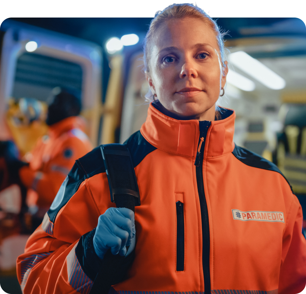 Eine lächelnde Rettungssanitäterin mit Rucksack und Hygienehandschuhen, im Hintergrund steht ein Rettungswagen mit geöffneten Heckklappen