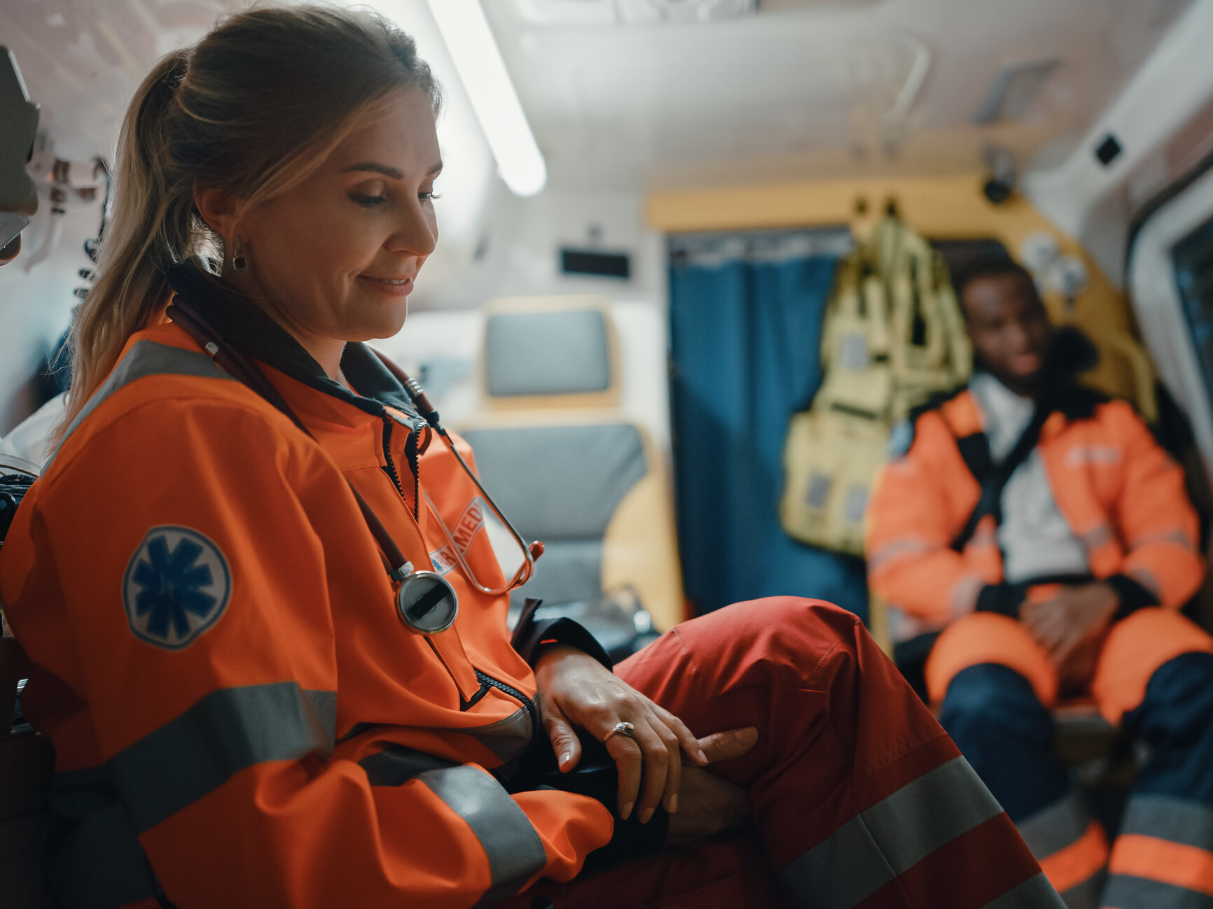 Vor sich hin lächelnde Rettungssanitäterin im Rettungswagen sitzend, im Hintergrund sitzt ein zweiter Rettungssanitäter.