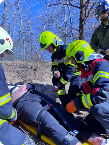 Draußen-Einsatz: Drei Personen in Einsatzkleidung der Feuerwehr knien neben einer Person, die auf einer Trage festgeschnallt ist.